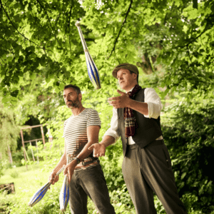 Interaktive Jonglage: Künstler und ein Erwachsener Teilnehmer üben geminsam mit Keulen im Sonnenschein.
