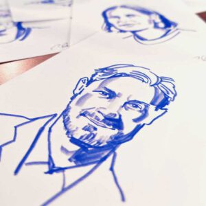 Drei fertige Live-Zeichnungen: Ein detailliertes Porträt im Fokus im Vordegrund, flankietrt von weiteren Zeichnungen im HIntergrund. Entstanden auf einer Messe in Nürnberg.