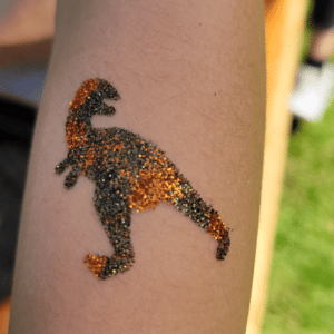 Glitzer-Tattoo eines Dinosaurier-Motivs in Braun und Grün auf dem Arm eines Kindes.